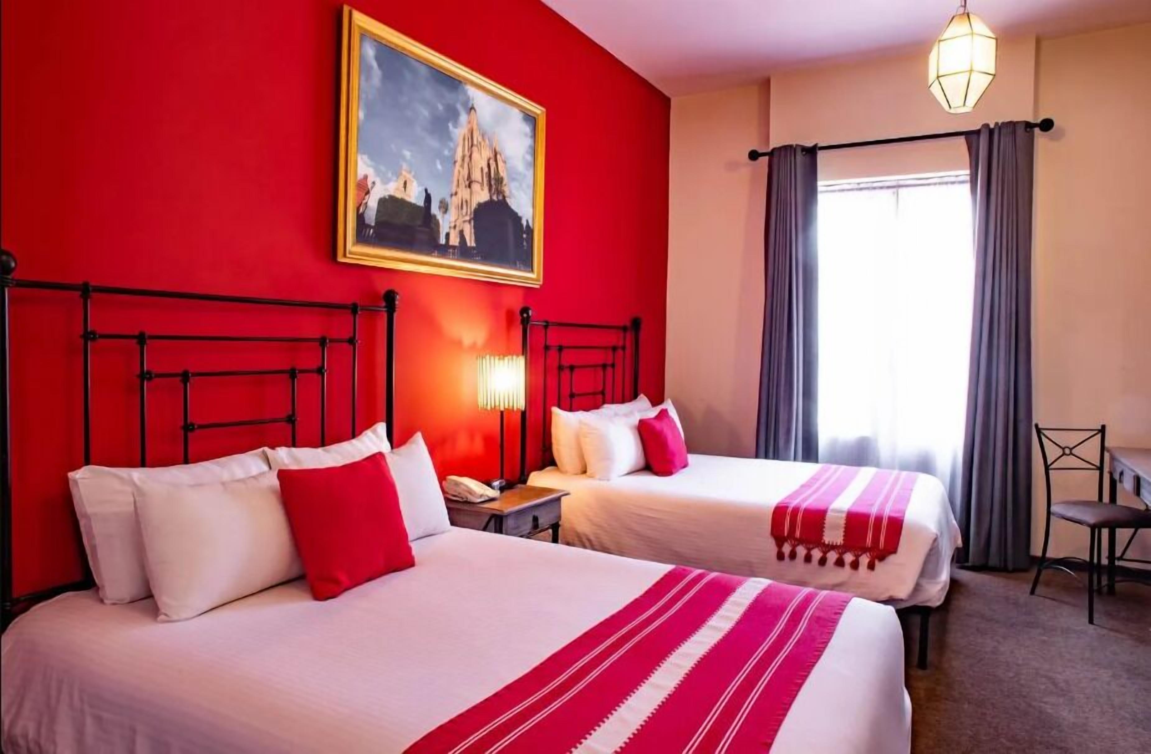 A hotel room with two beds, red walls, a painting of a cathedral, and a window with curtains.
