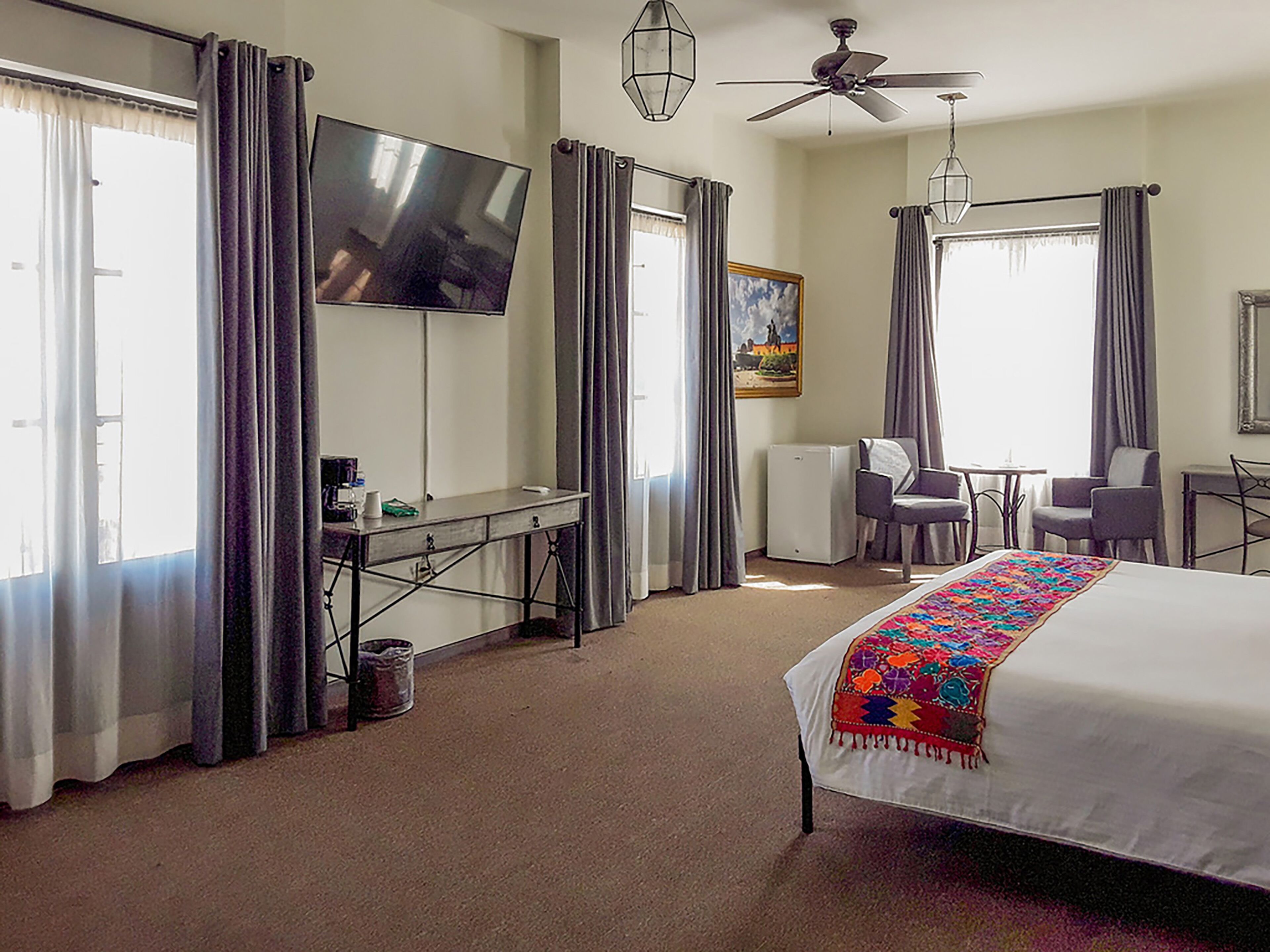 A hotel room with a bed, desk, chairs, a TV, and a ceiling fan.
