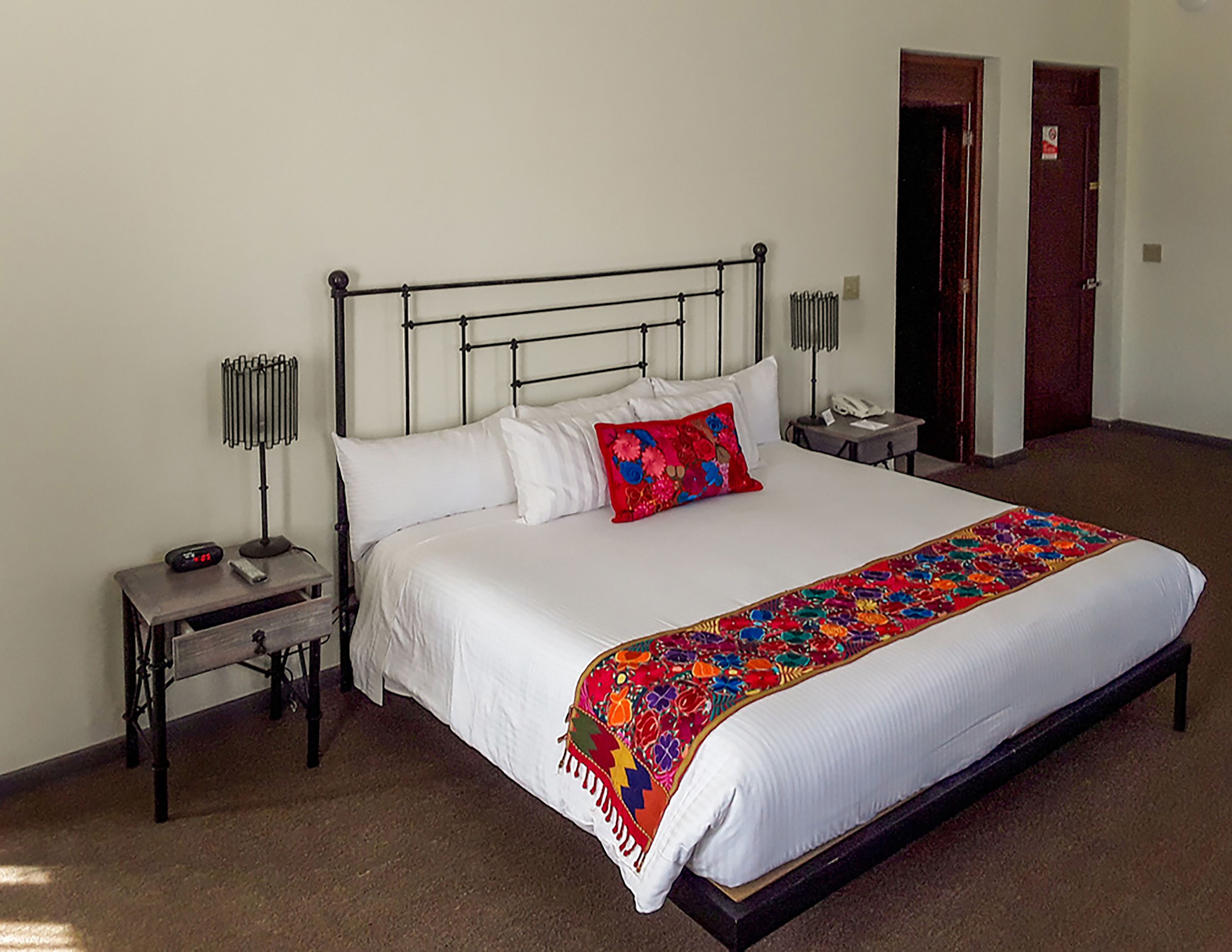 A neatly made bed with white linens, a colorful throw blanket, and a red pillow. There is a bedside table with a lamp and a phone. The room has a simple, clean design with a carpeted floor.