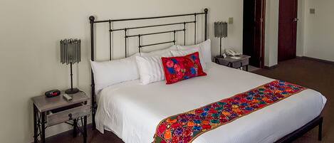 A neatly made bed with white linens, a colorful throw blanket, and a red pillow. There is a bedside table with a lamp and a phone. The room has a simple, clean design with a carpeted floor.