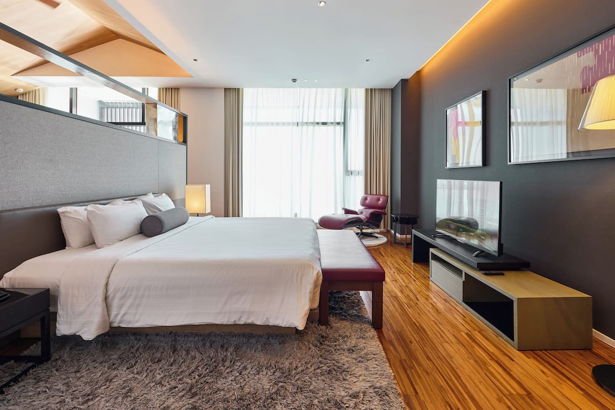 A modern hotel room with a large bed, a flat-screen TV, a seating area, and a view of the city through the window.