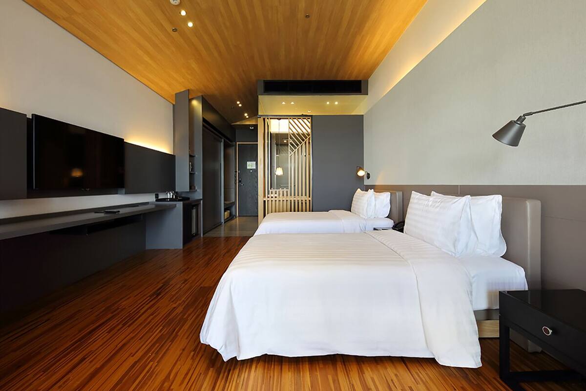 A modern hotel room with a wooden floor, a large bed with white bedding, a flat-screen TV mounted on the wall, and a bedside table with a lamp.