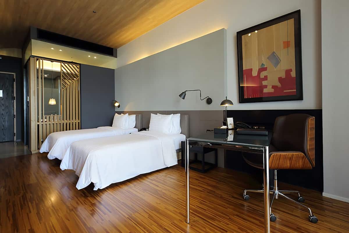 A hotel room with two beds, a desk, a chair, a lamp, and a painting on the wall.