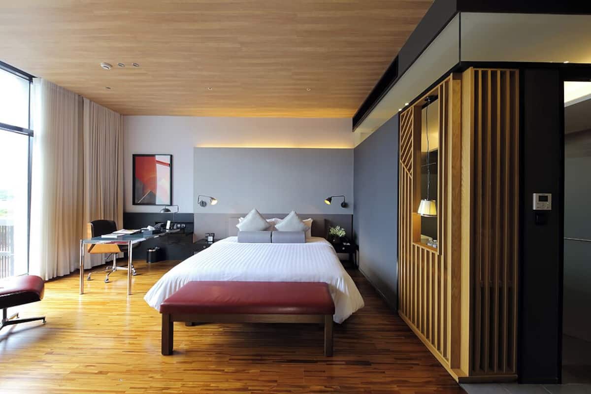 A modern hotel room with a large bed, a desk, and a chair. There is a bench, a lamp, and a wall-mounted TV.