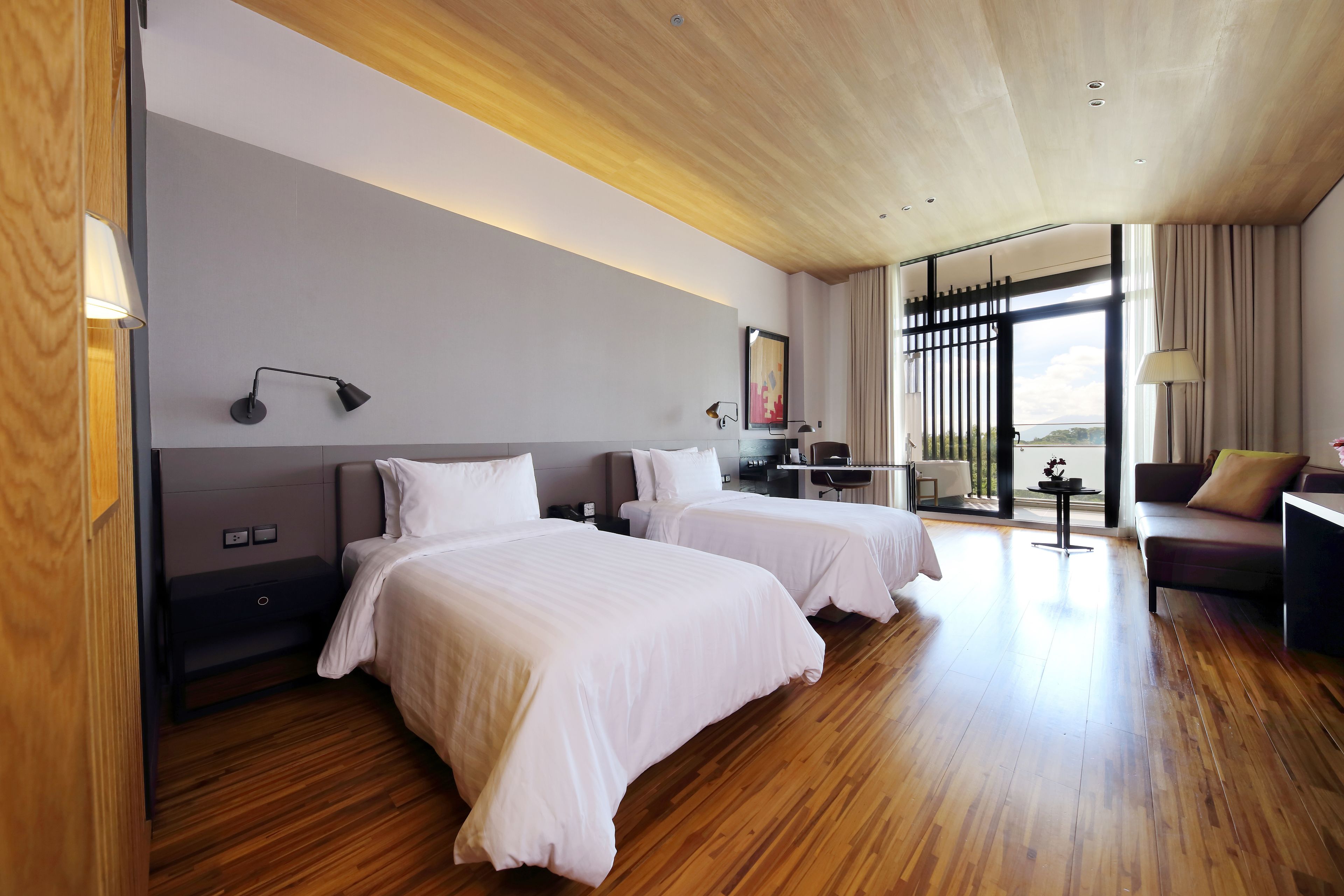 A hotel room with two beds, a desk, a sofa, and a large window with curtains.