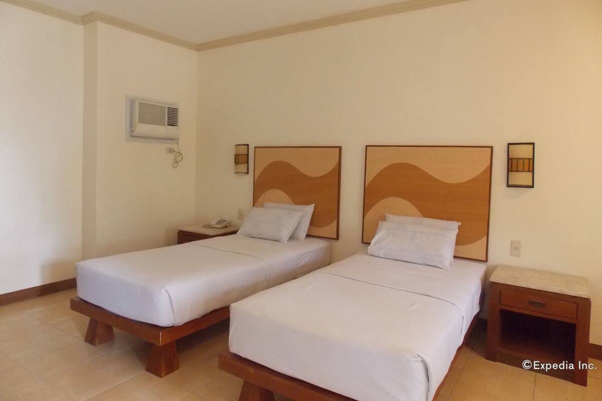 A hotel room with two single beds, a bedside table with a lamp, a wall-mounted air conditioner, and a small wooden nightstand.