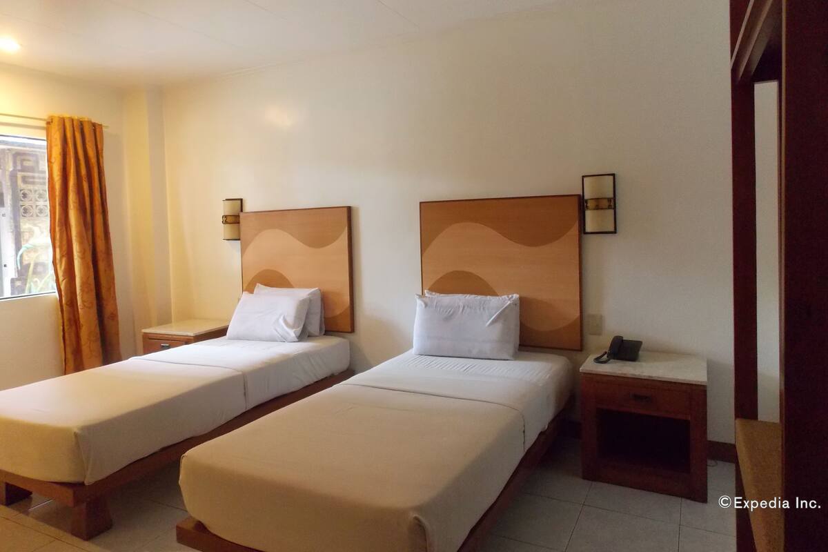 A hotel room with two single beds, a nightstand with a phone, a wall-mounted lamp, and a window with curtains.