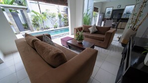 A modern living room with two brown sofas, a coffee table with a flower vase, and a TV. There is a pool visible through the glass doors. - Bahagia Villas (Denpasar)