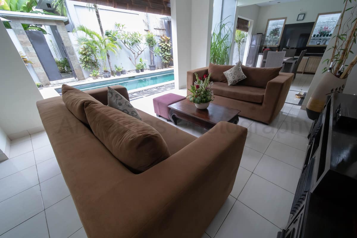 A modern living room with two brown sofas, a coffee table with a flower vase, and a TV. There is a pool visible through the glass doors.