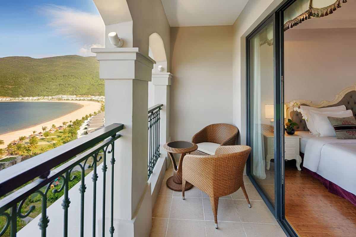 A balcony with a view of a beach and mountains, a bed with white bedding, and wicker furniture.