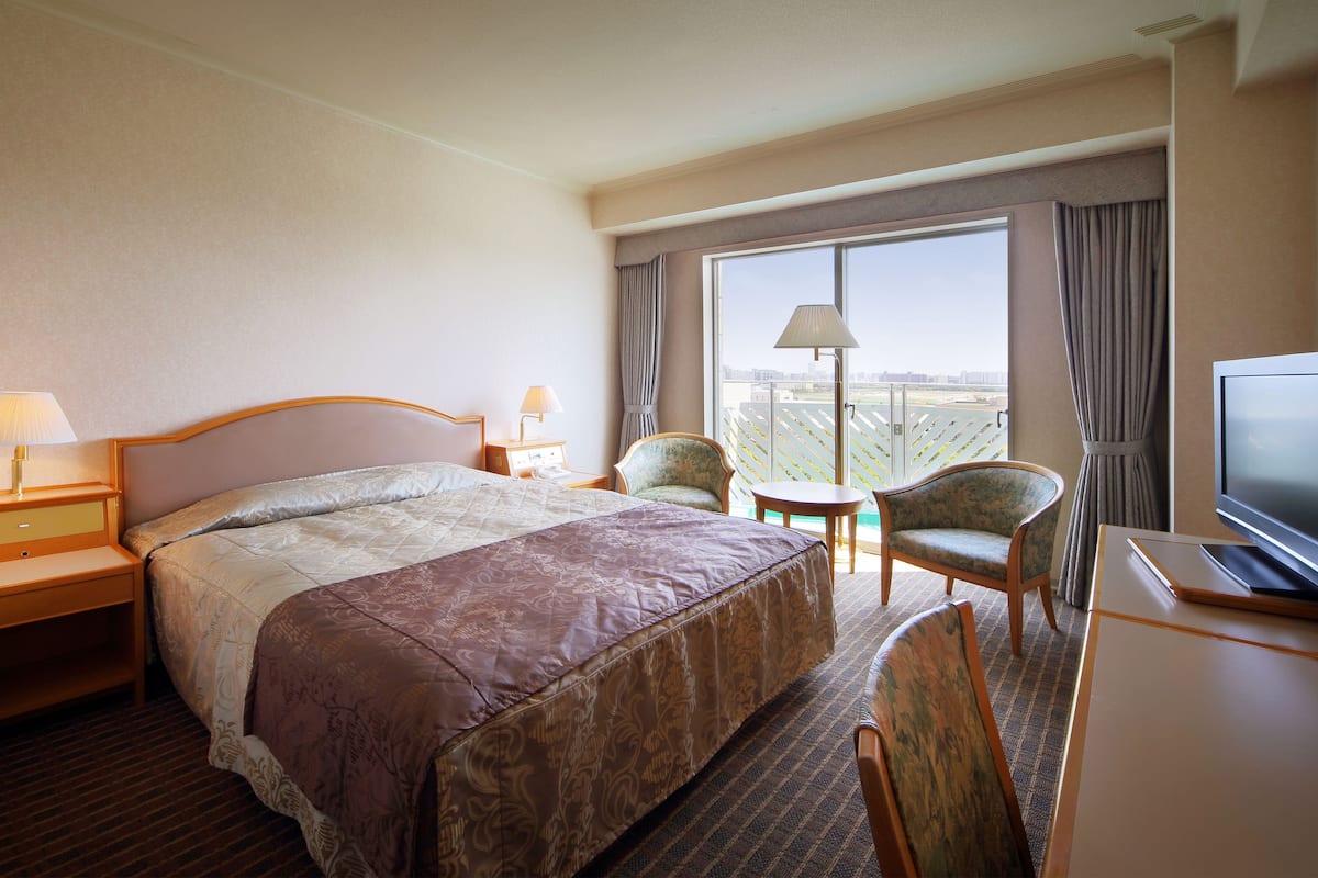A hotel room with a large bed, a desk with a TV, a chair, a lamp, and a view of the city through the window.