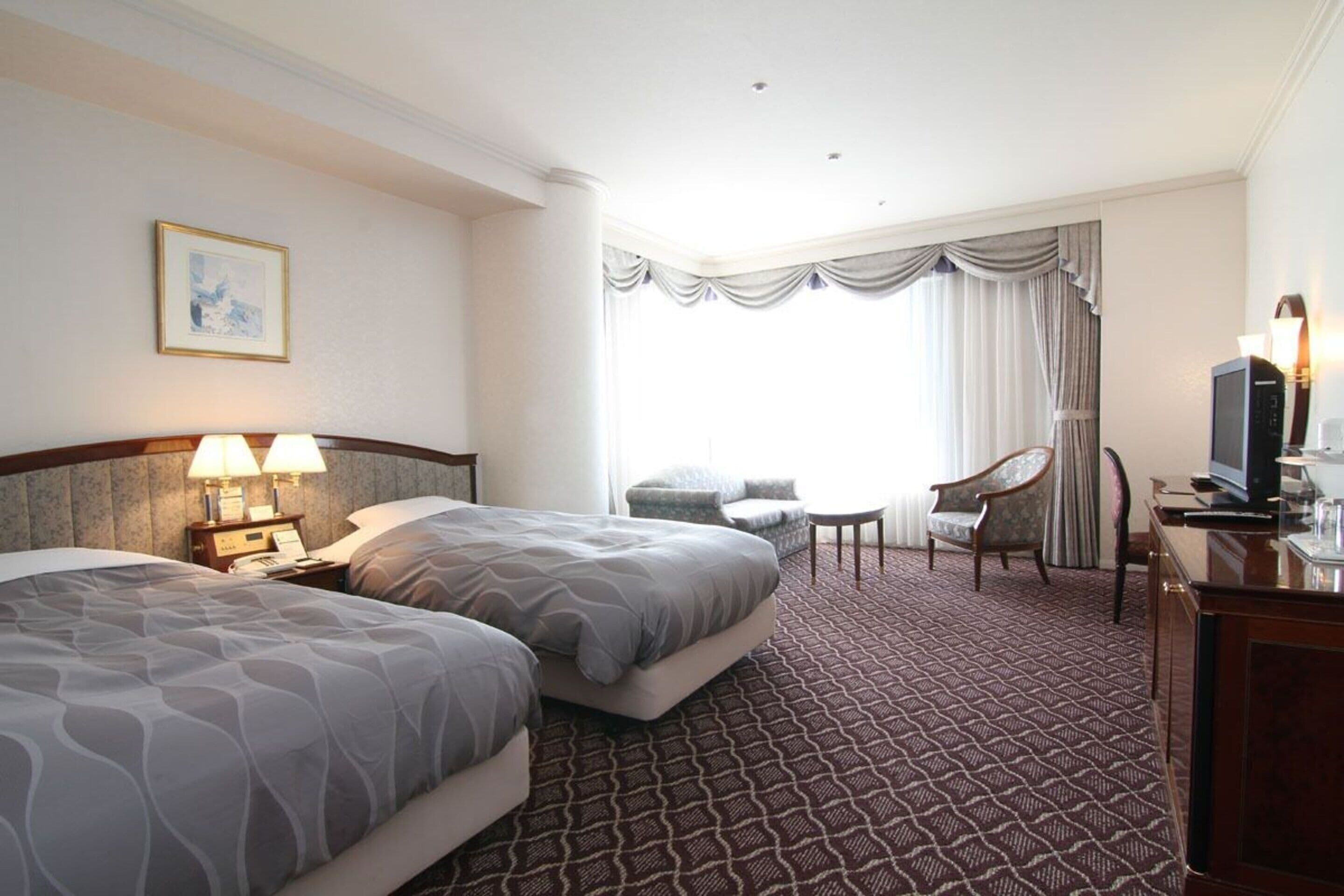 A hotel room with two beds, a desk with a television, a chair, and a window with curtains.