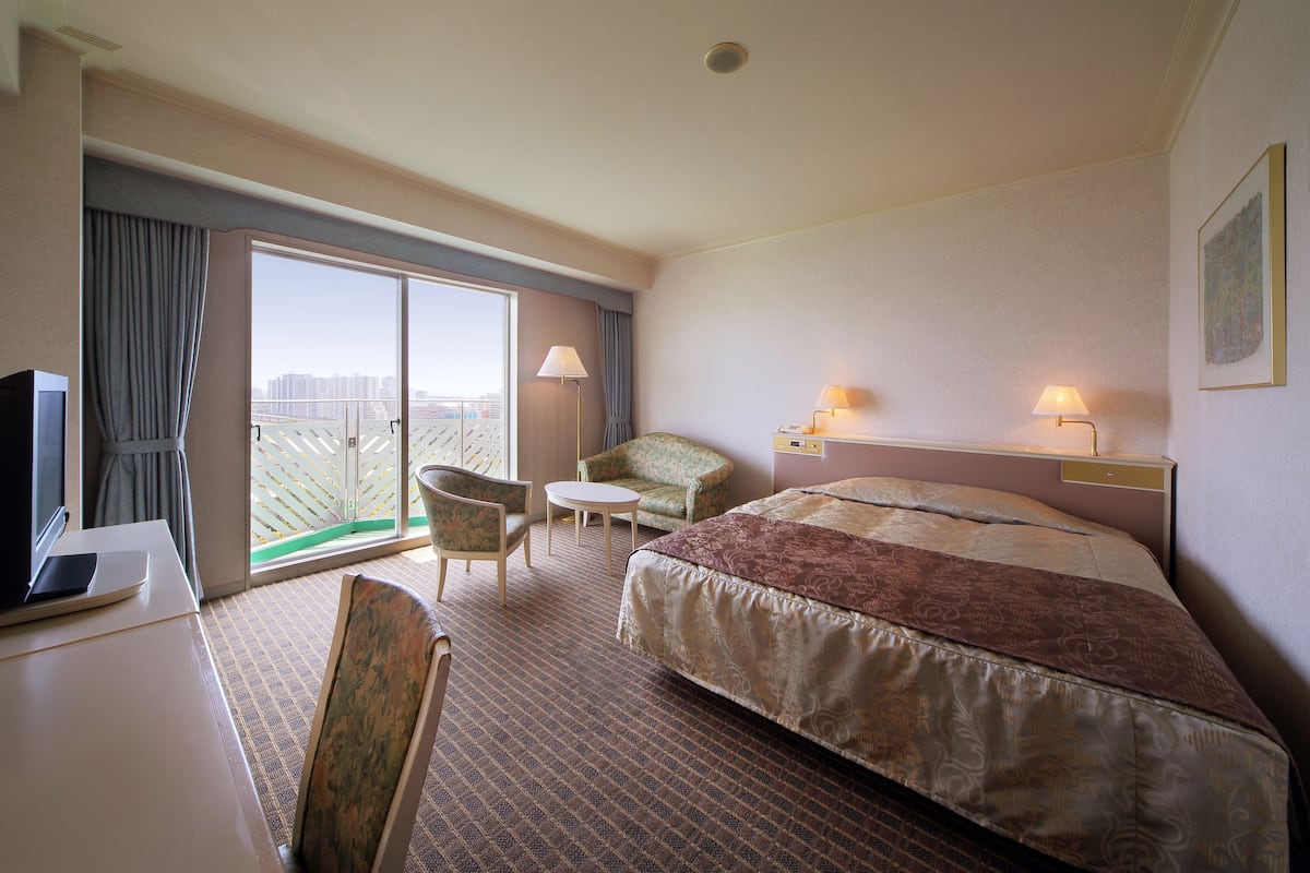 A hotel room with a bed, a desk with a laptop, a chair, a small table, and a view of the city through the window.