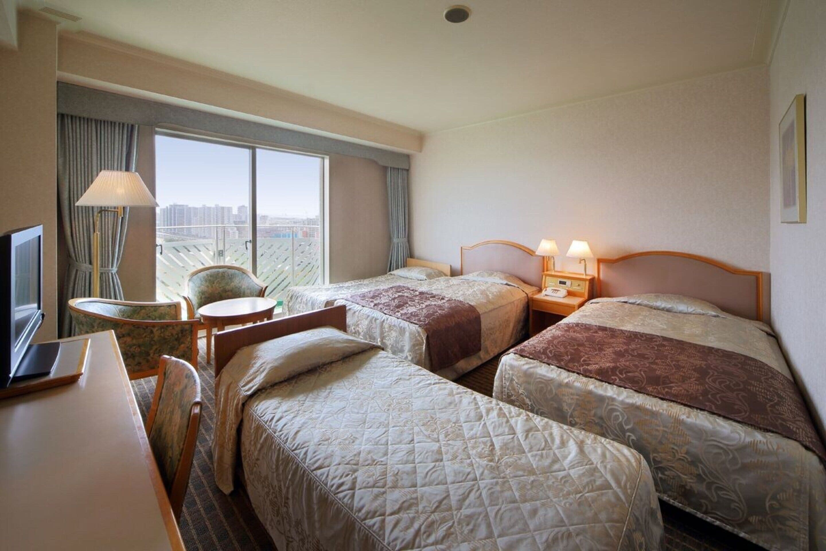 Een hotelkamer met twee bedden, een bureau met een laptop, een stoel, een lamp en uitzicht op de stad door grote ramen.
