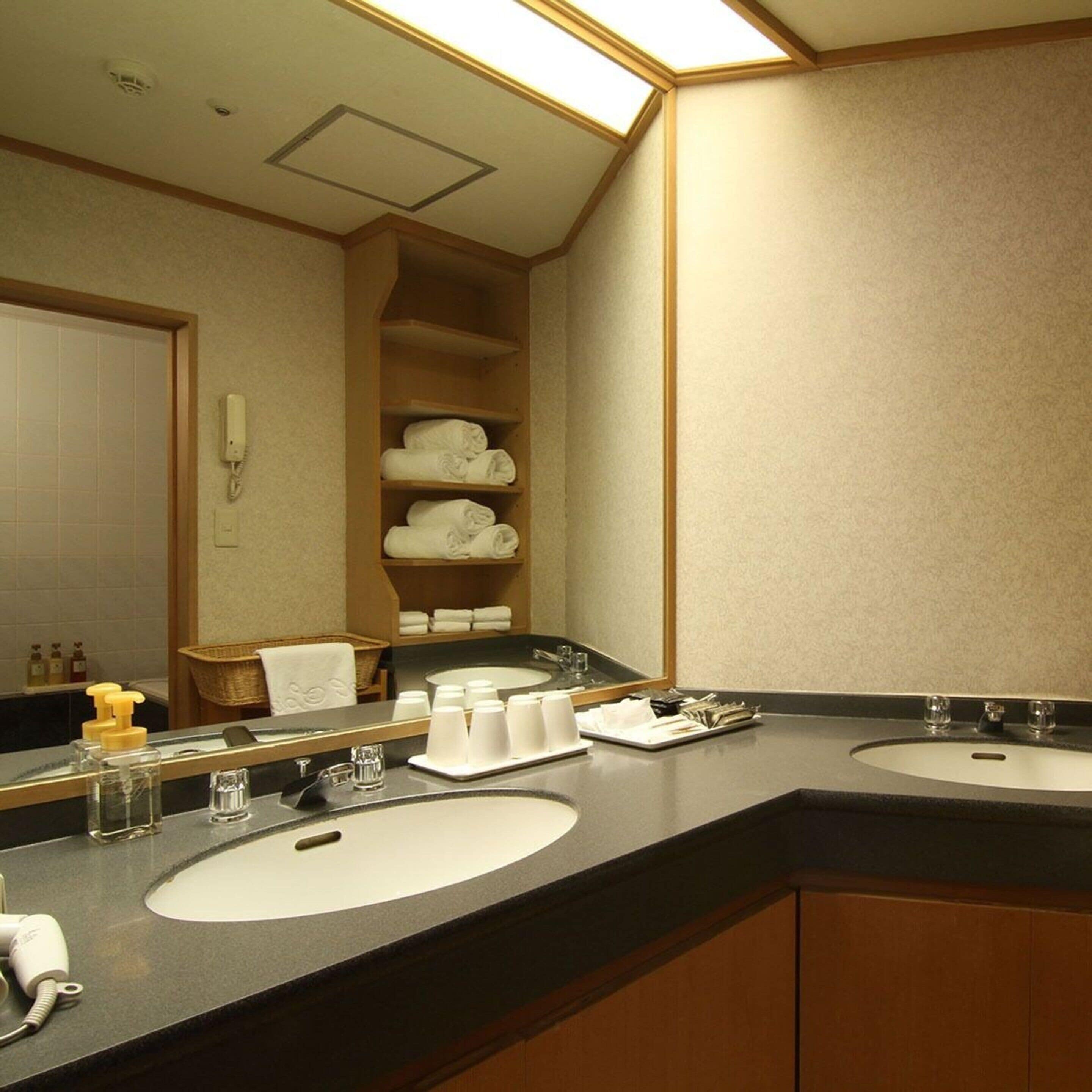 A hotel bathroom with two sinks, a mirror, a towel rack, and a shelf with folded towels.