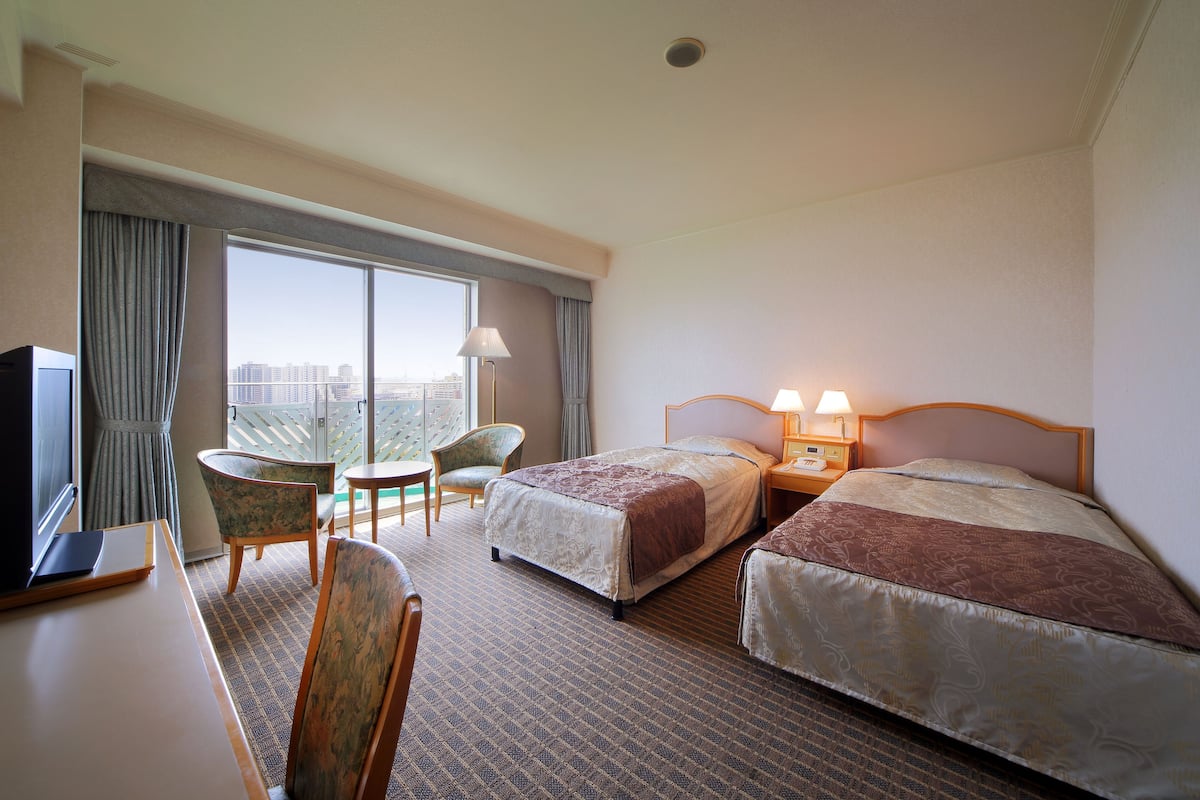 A hotel room with two beds, a desk with a TV, a chair, a lamp, and a view of the city.
