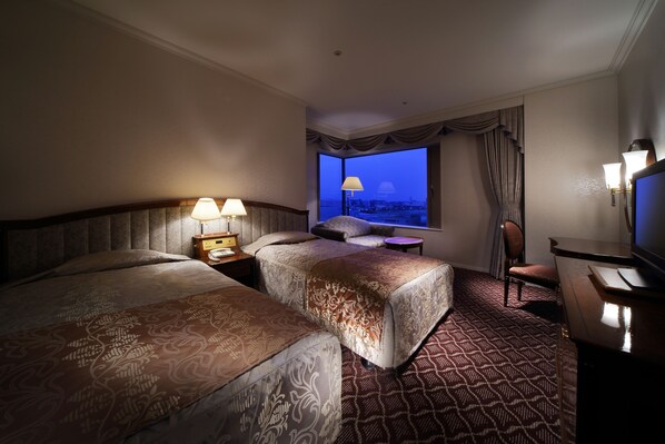 A hotel room with two beds, a desk, a TV, and a window with a city view.