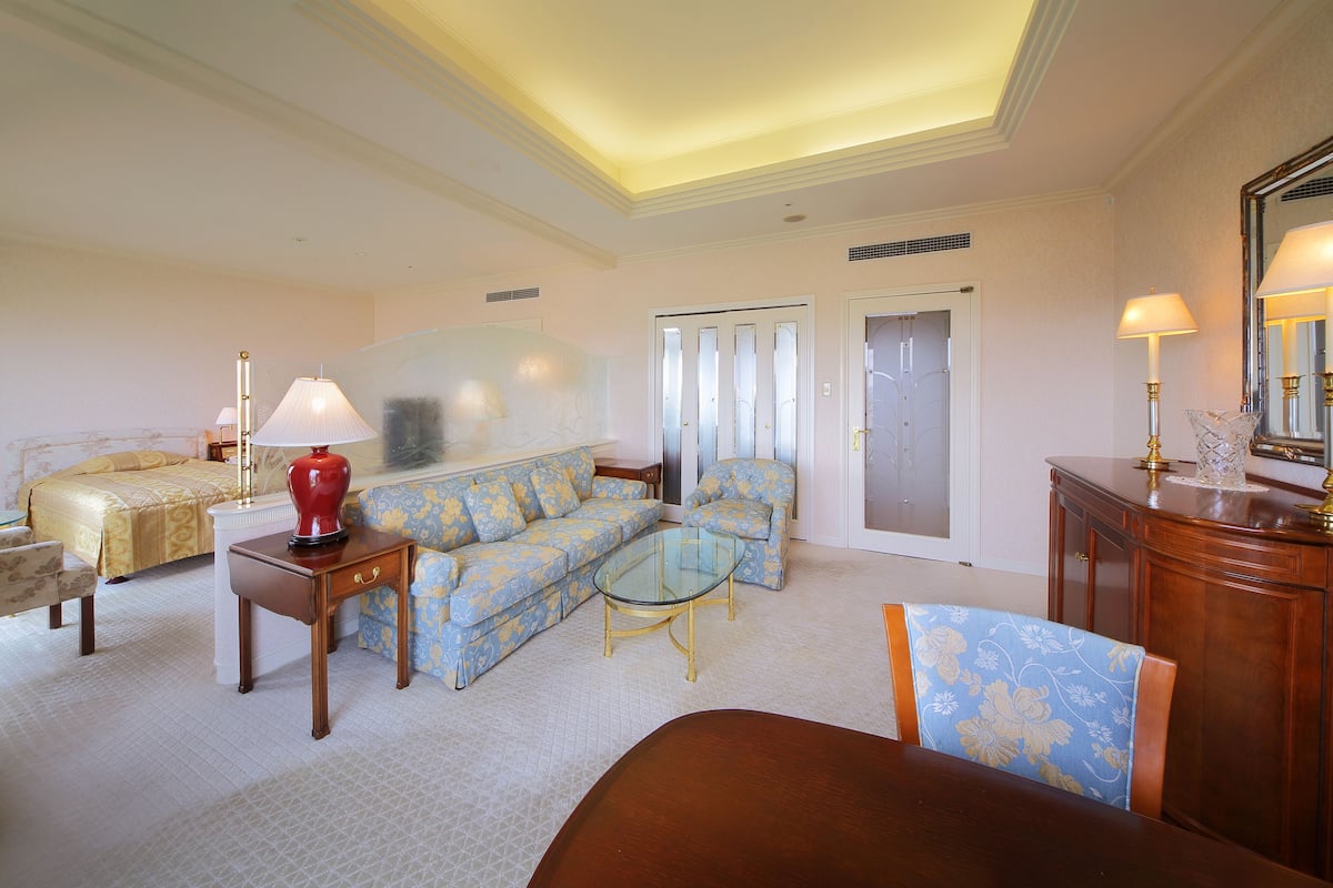 A hotel room with a bed, a sofa, a glass coffee table, a wooden side table, and a desk with a lamp.