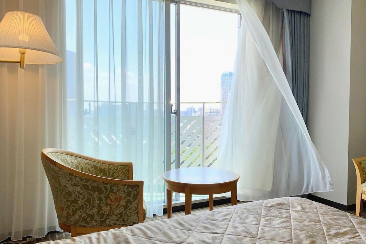 Habitación de hotel con cama, una silla, una mesita y un ventanal con cortinas.