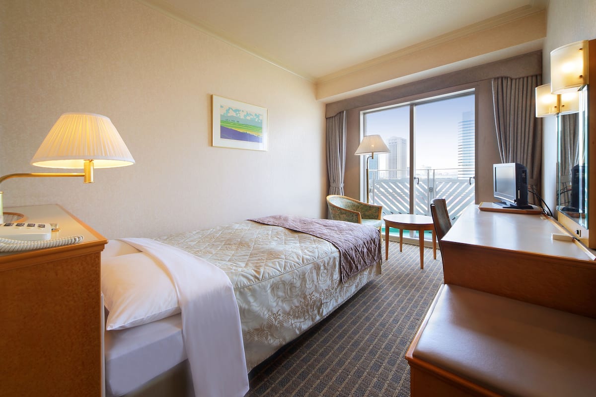 A hotel room with a bed, a desk, a chair, a desk lamp, a telephone, and a window with a view of a building.