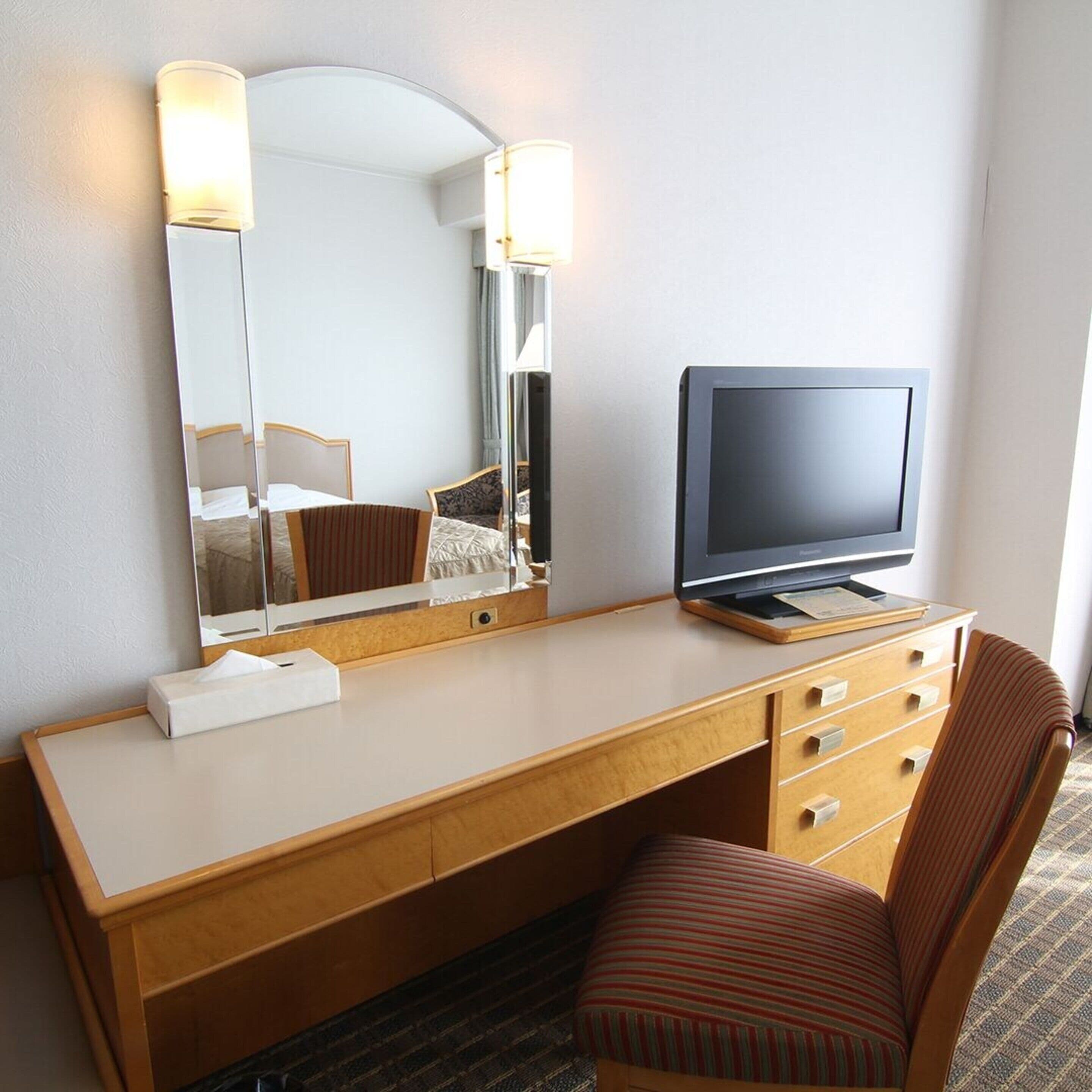 A hotel room with a wooden desk, a television, a mirror, and a striped chair.