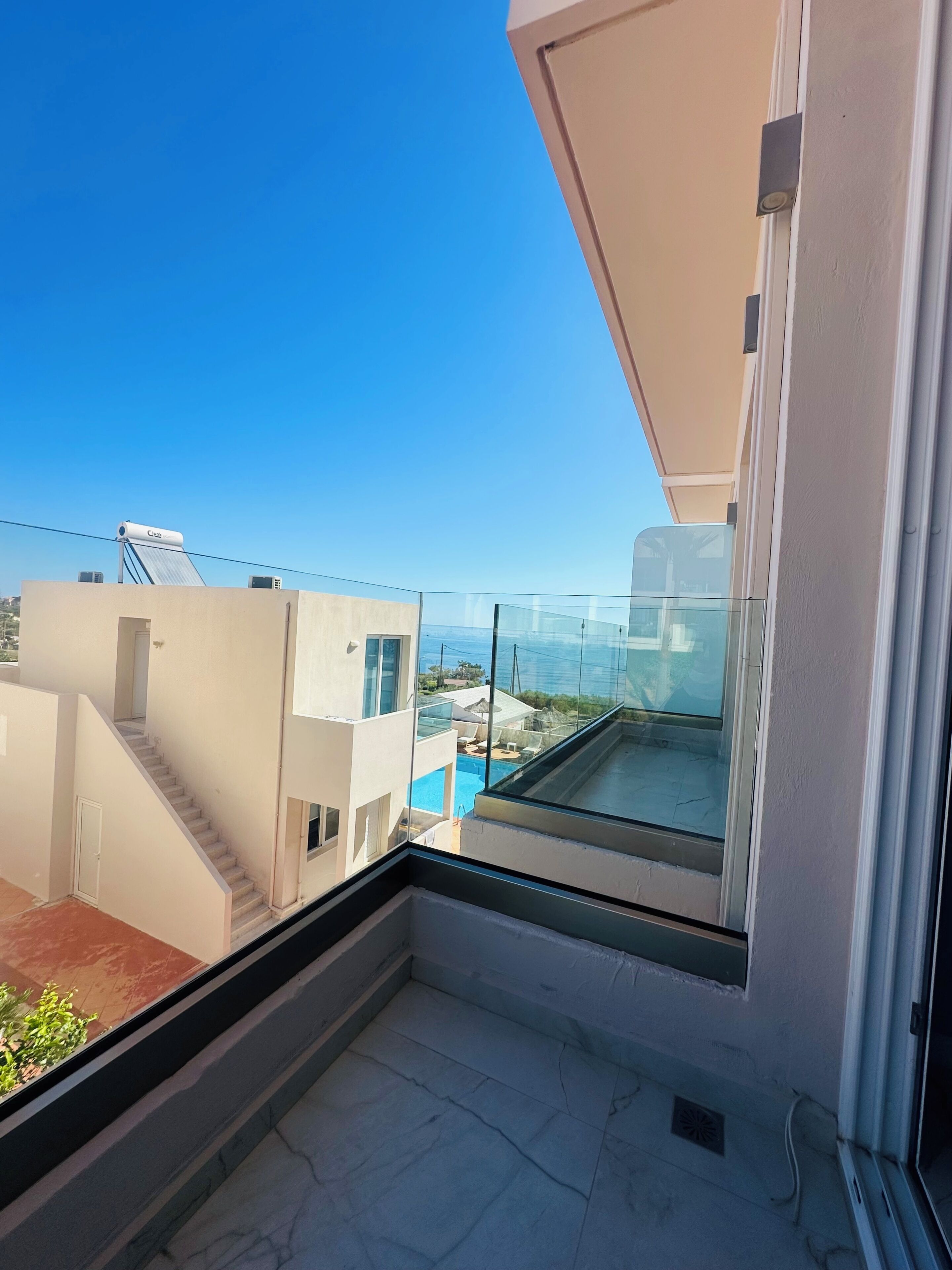 A balcony with a glass railing, a clear blue sky, and a view of a swimming pool and neighboring buildings.