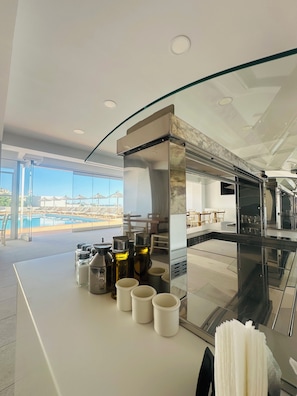 Cocina moderna con separador de vidrio, vista a la piscina y comedor con mesas y sillas.