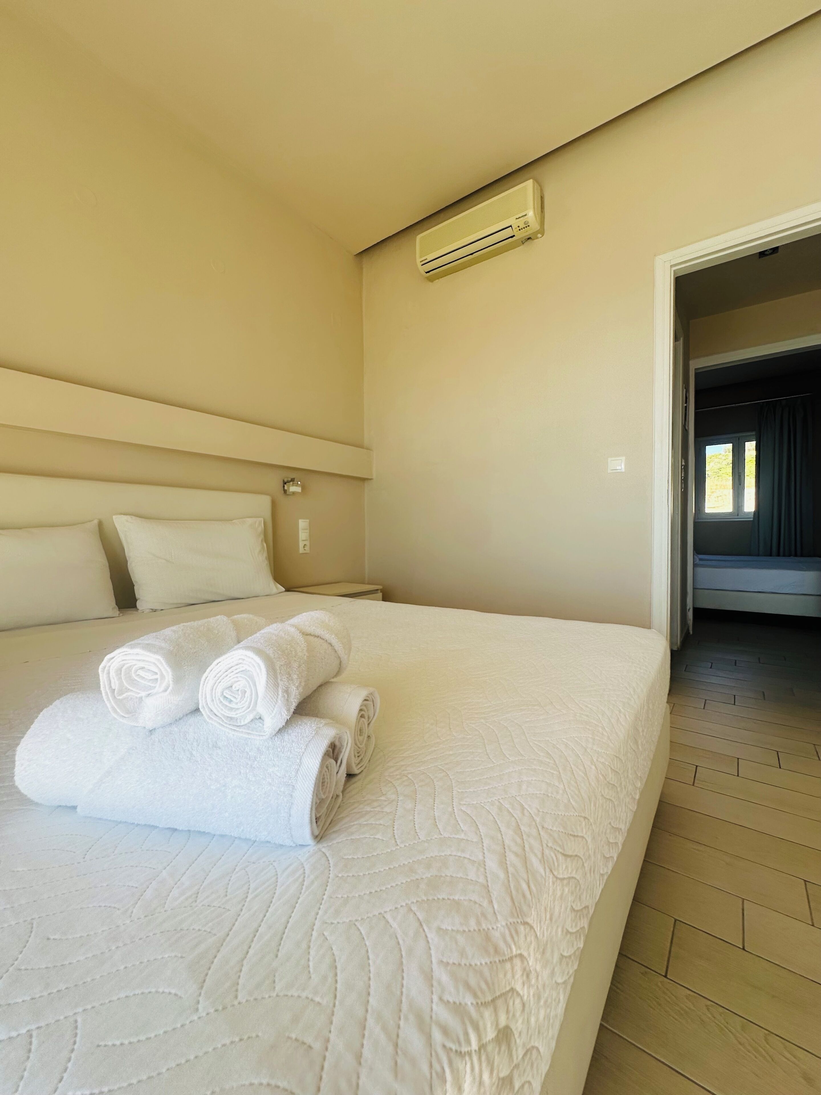 Ein Hotelzimmer mit Bett, einer Wandklimaanlage und Blick auf ein anderes Zimmer durch eine offene Tür.