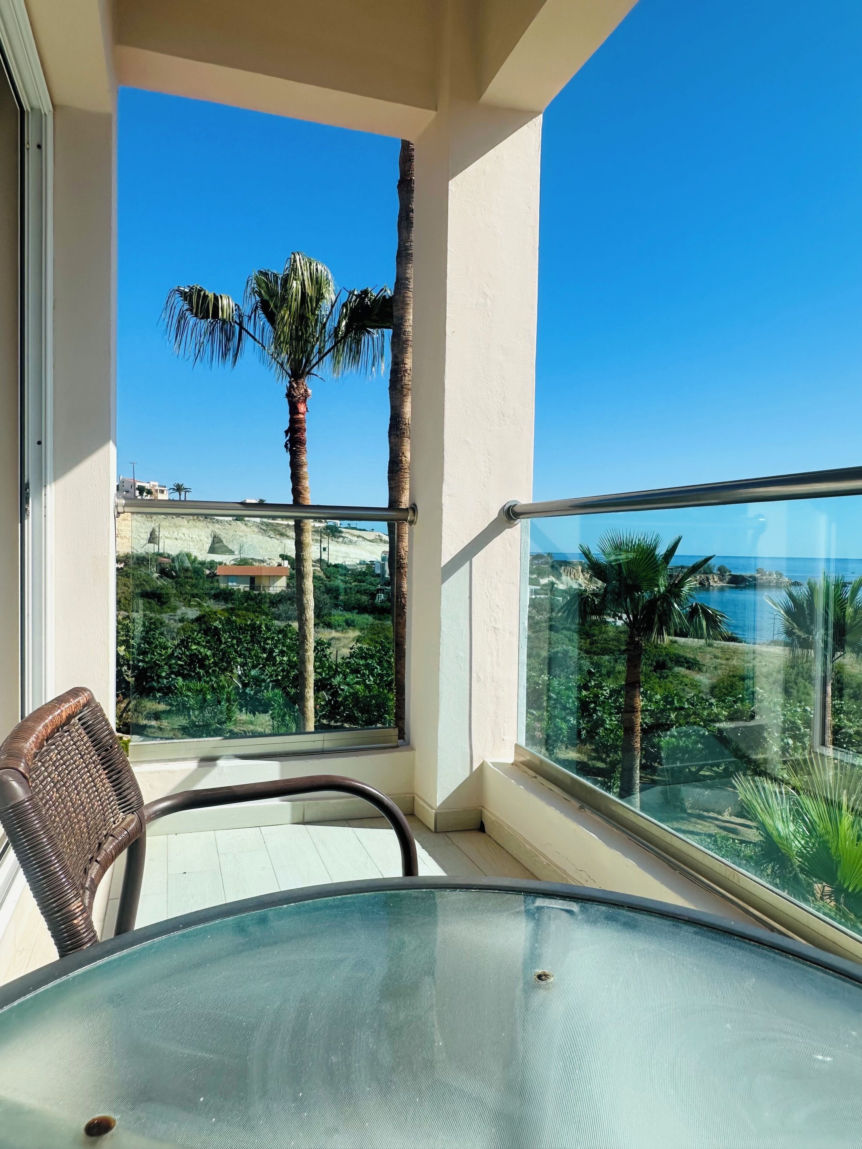 Een balkon met een glazen tafel en stoelen, met uitzicht op een kustlandschap met palmbomen en een helderblauwe lucht.