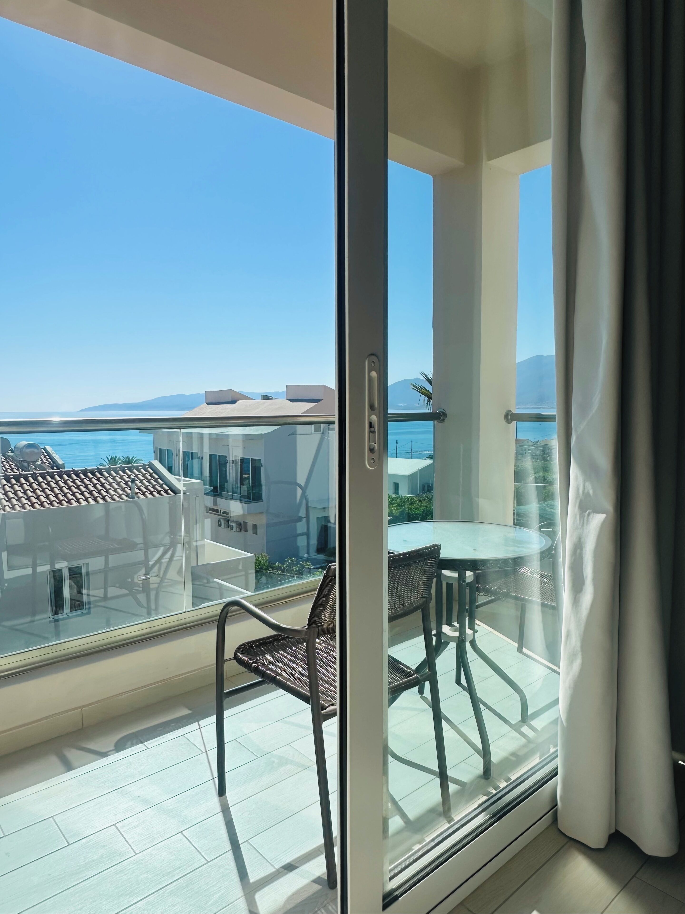 A balcony with a glass door, a table with chairs, and a view of the sea and buildings.