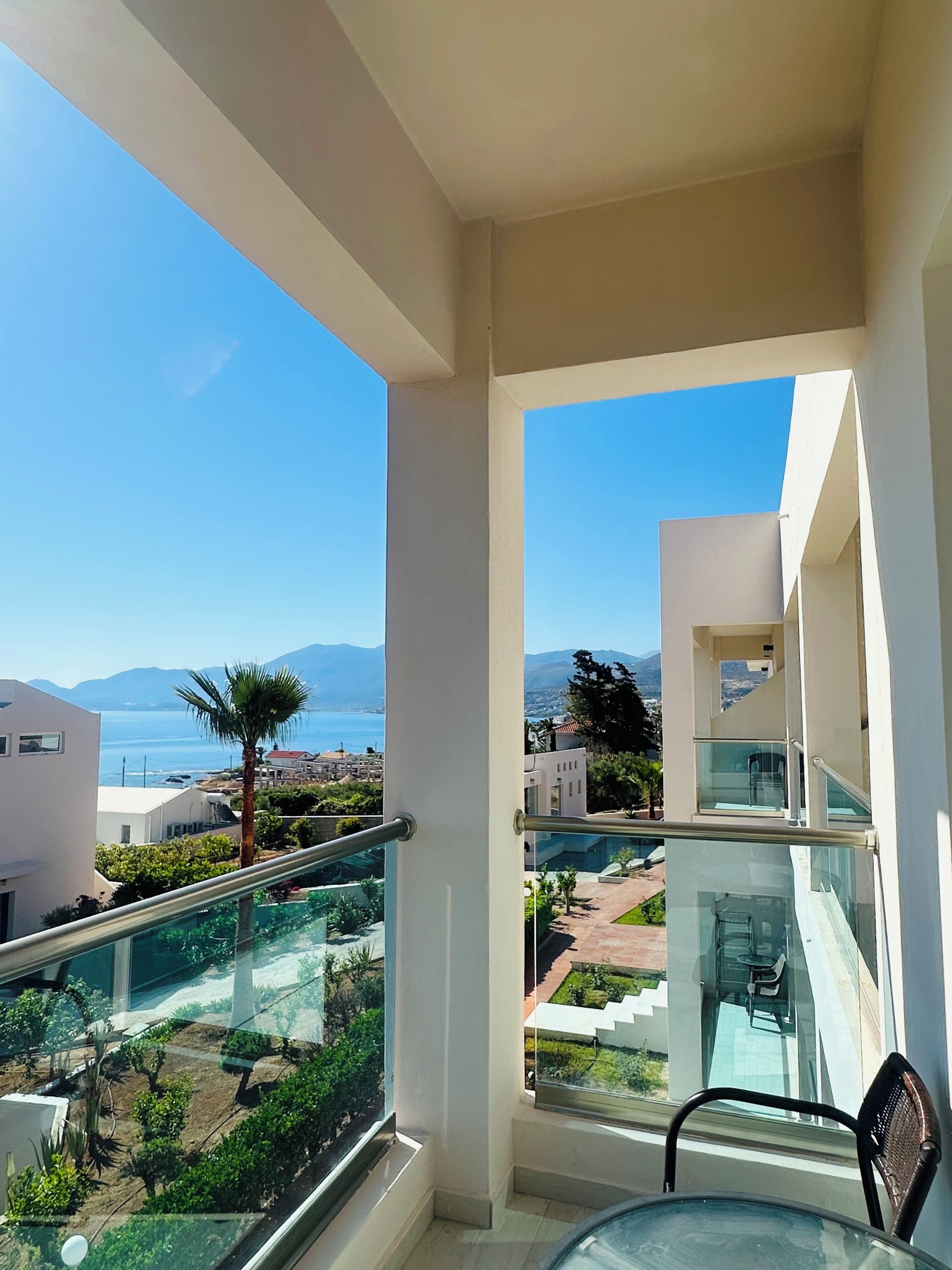 A balcony with a glass railing, a chair, and a view of the sea and mountains.