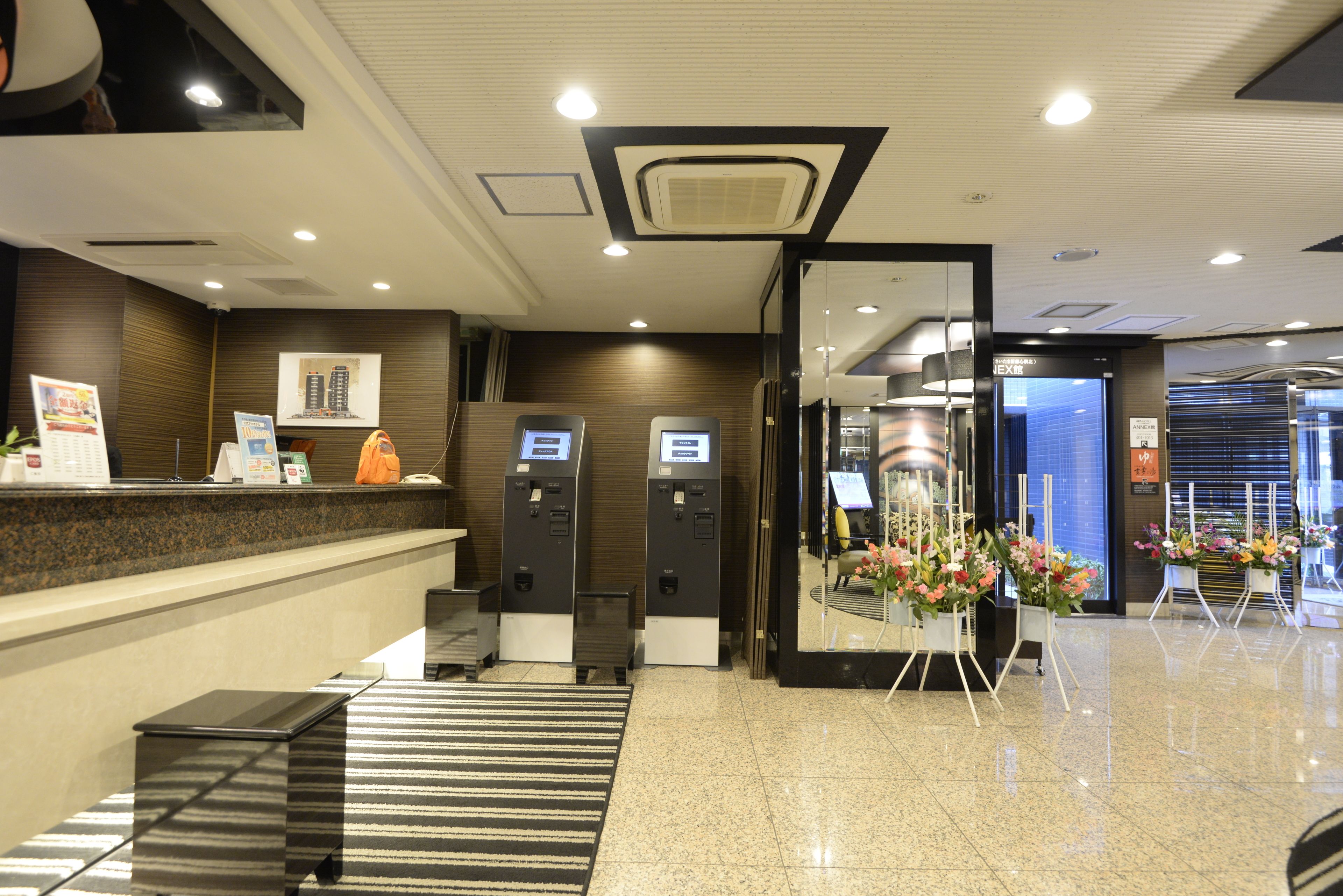 A hotel reception area with a reception desk, two self-service kiosks, a check-in counter, and a floral display.