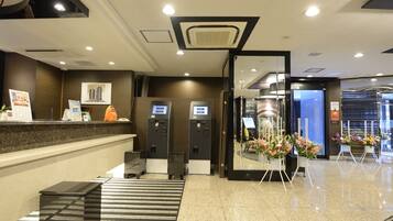 A hotel reception area with a reception desk, two self-service kiosks, a check-in counter, and a floral display.