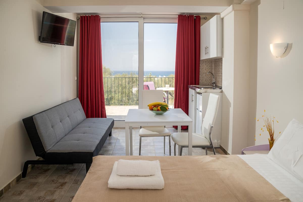 A hotel room with a bed, a sofa, a dining table with chairs, a TV mounted on the wall, and a balcony with red curtains.