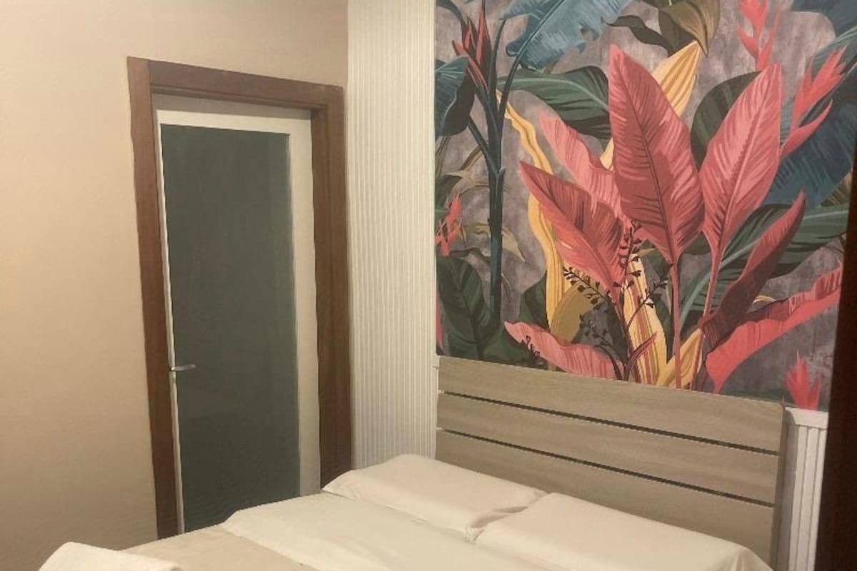 A hotel room with a bed, a door, and a large tropical plant mural on the wall.