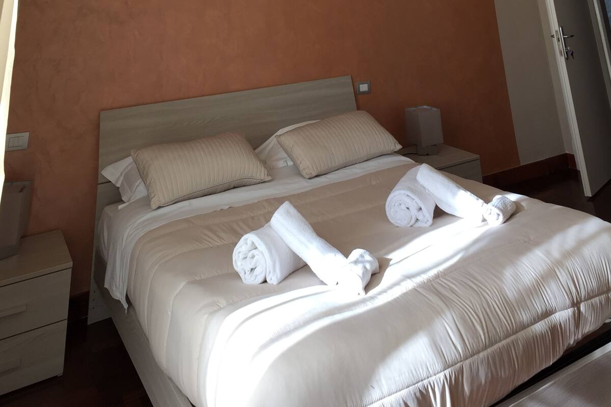 A neatly made bed with white linens and two rolled towels, a bedside table with a lamp, and a wooden headboard.