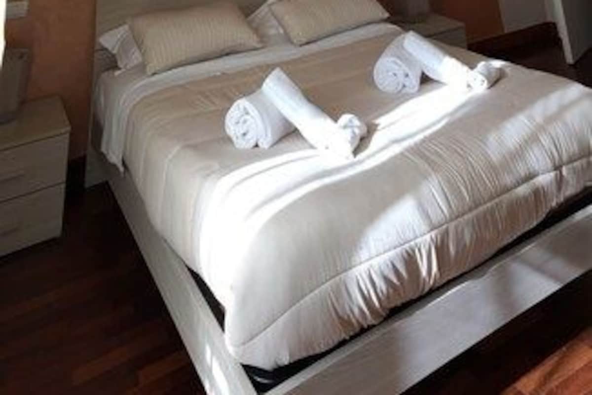 Un letto rifatto con asciugamani arrotolati, un comodino e un tavolino di fianco al letto in una stanza con pavimento in legno e parete spoglia.