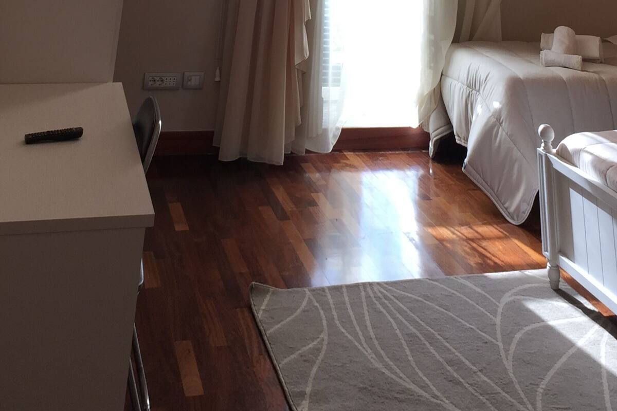 A hotel room with a wooden floor, a bed with white bedding, a desk with a remote control, and a window with beige curtains.