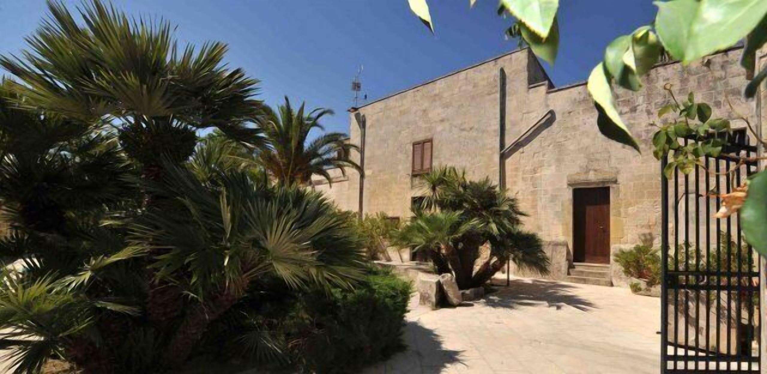 Un cortile con un edificio in pietra, palme e un cancello in metallo.