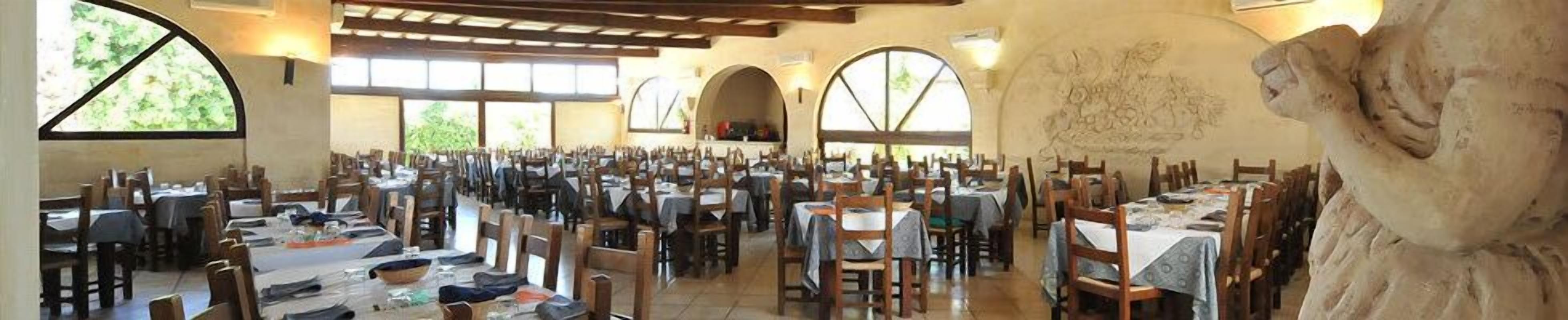 A large dining hall with multiple tables and chairs, arched windows, and a rustic interior.