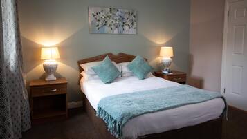 A neatly made bed with a blue blanket and pillows, flanked by two bedside tables with lamps, in a room with a light blue wall and a door to the right.