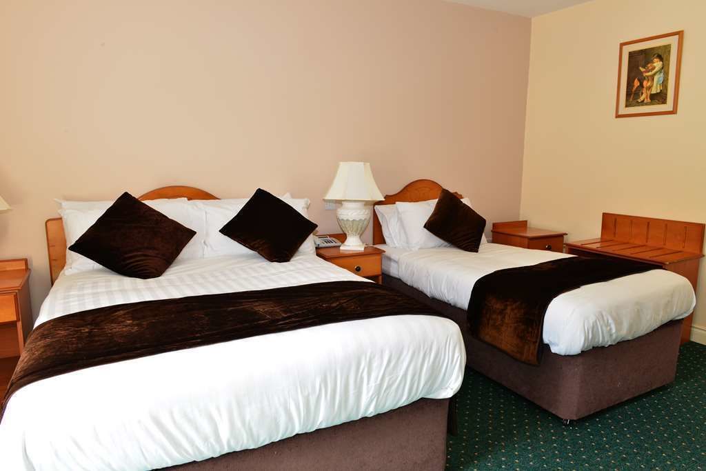 Habitación de hotel con dos camas, cada una con cabecero de madera, ropa de cama blanca y mantas marrones oscuras. Hay una mesita de noche con lámpara y una pequeña mesa entre las camas.