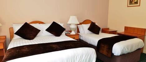 Habitación de hotel con dos camas, cada una con cabecero de madera, ropa de cama blanca y mantas marrones oscuras. Hay una mesita de noche con lámpara y una pequeña mesa entre las camas.