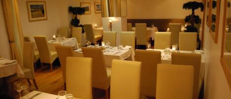 Comedor con varias mesas dispuestas para dos comensales, cada una con mantel blanco, velas y cubiertos.