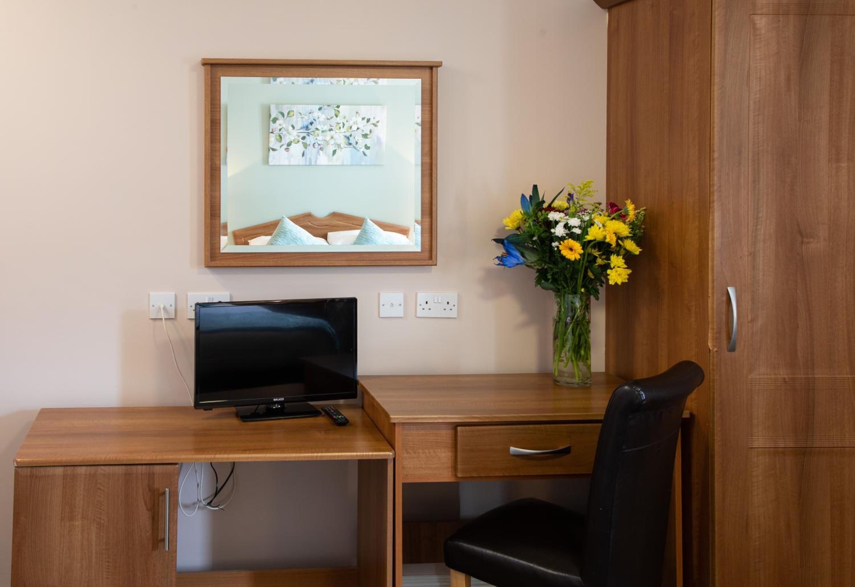 A wooden desk with a television, a vase of flowers, and a mirror on the wall.