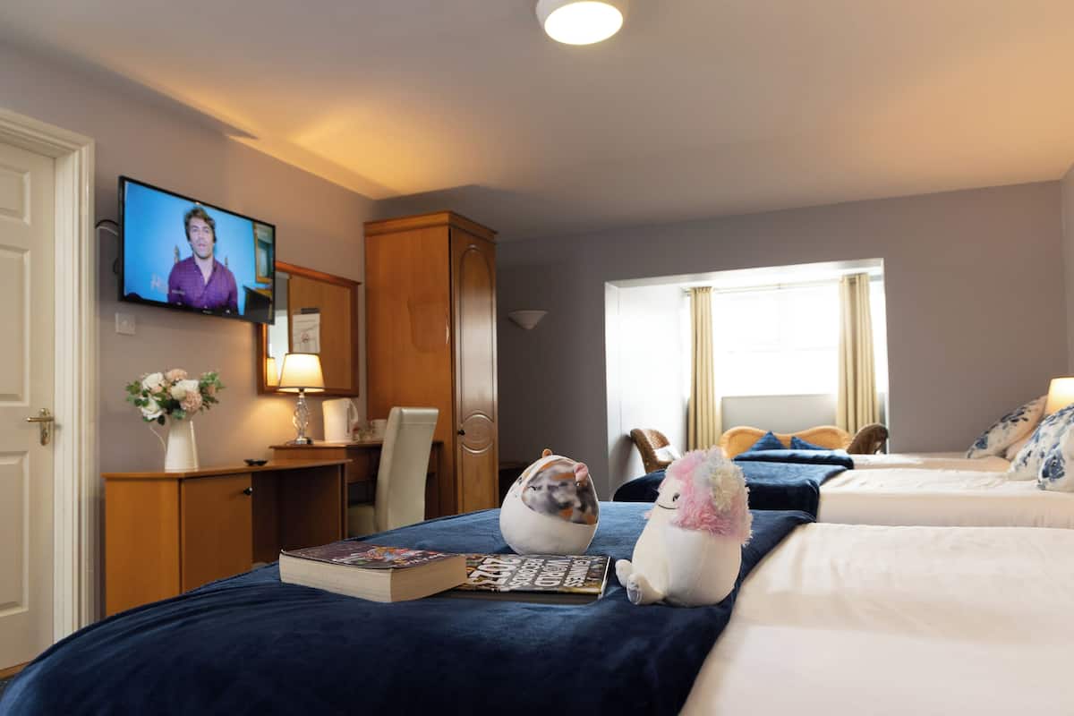 A hotel room with a TV on the wall, a desk with a lamp, a chair, a bed with a blue bedspread, a stuffed unicorn, and a book.