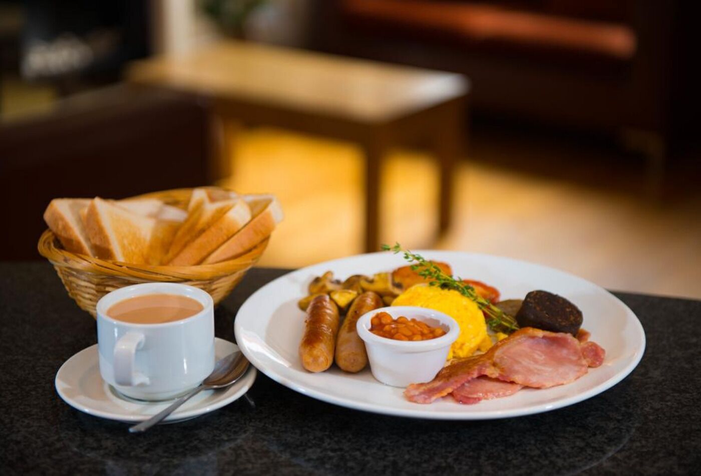 A breakfast plate with sausages, eggs, ham, and a small bowl of beans, accompanied by a cup of coffee and a basket of bread.