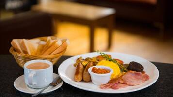 Prato de café da manhã com salsichas, ovos, presunto e uma pequena tigela de feijão, acompanhado de uma xícara de café e uma cesta de pão.