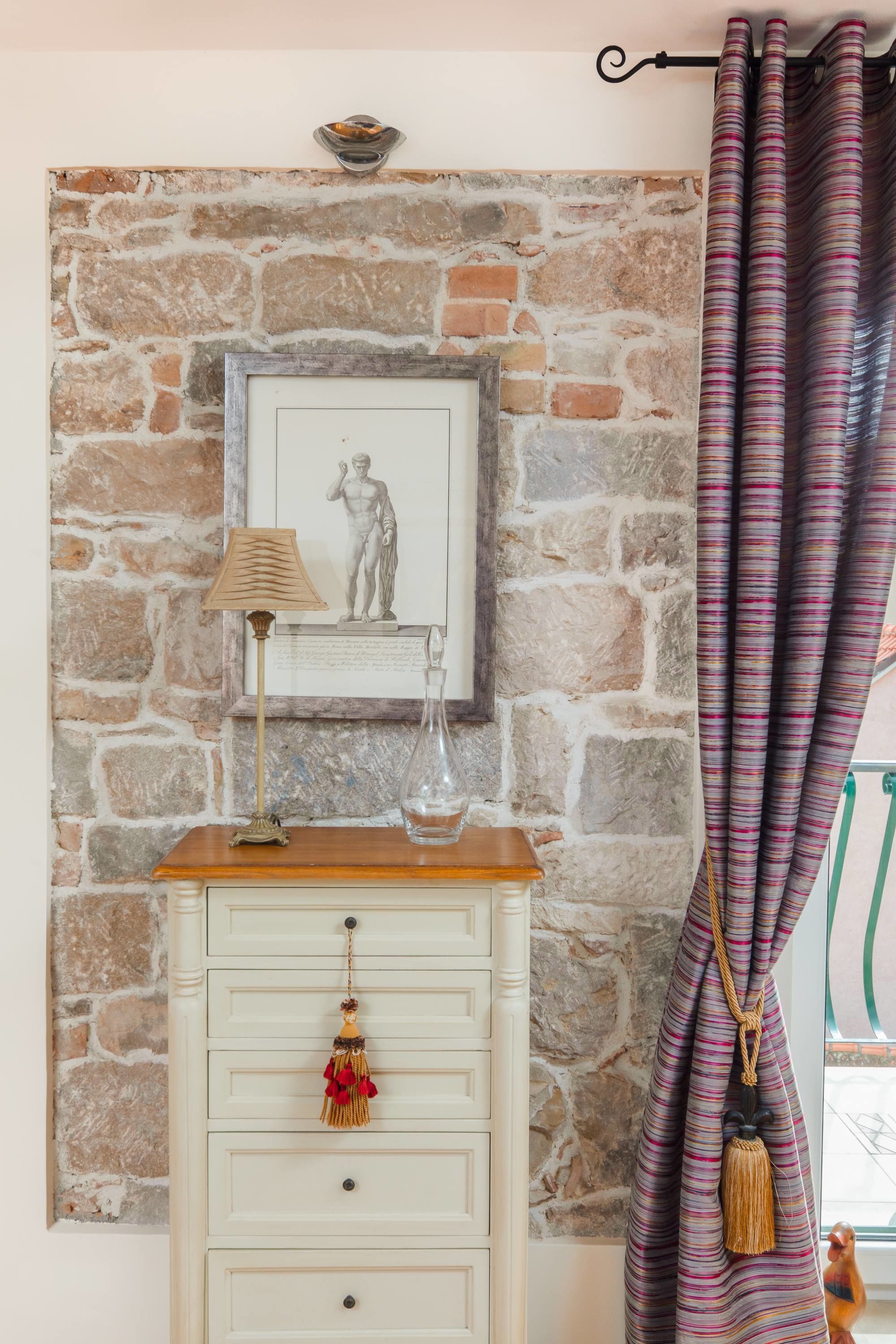 A room with a stone wall, a framed picture, a lamp, a small table, and a curtain.