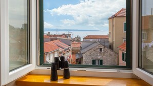 Una ventana con binoculares sobre un repisa de madera, con vista a un pueblo costero de edificios con tejados rojos y un cielo despejado.